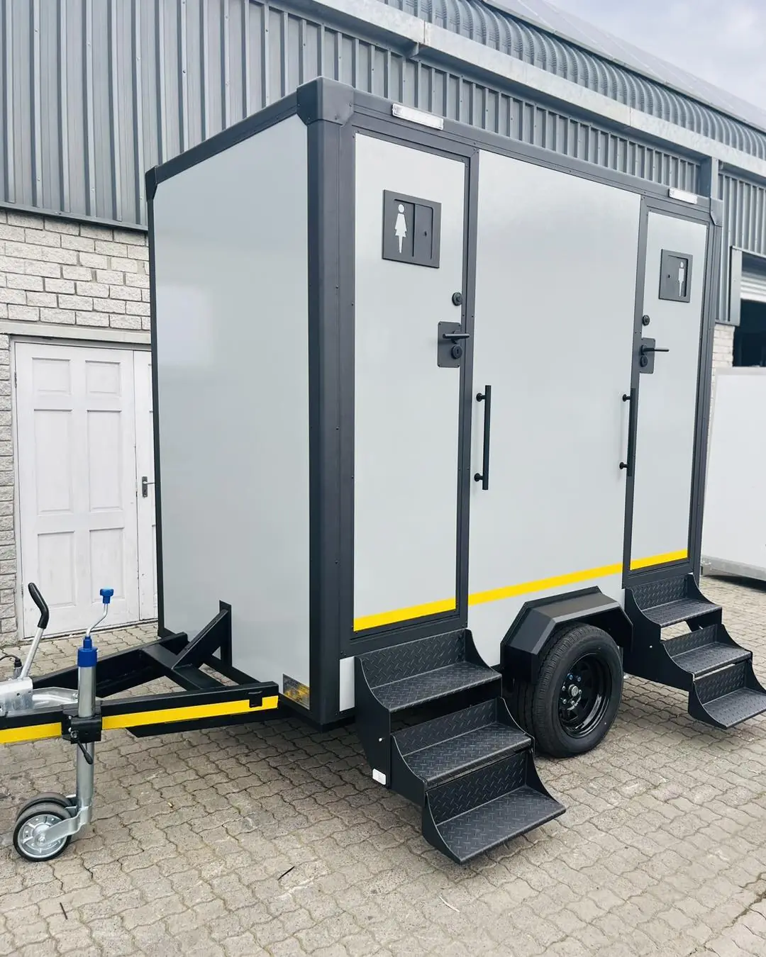 White two-station restroom trailer with black trim and fold-down steps on pavers